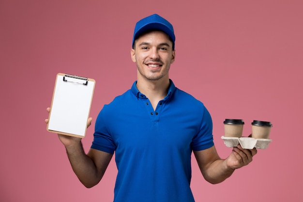 male courier in blue uniform holding delivery coffee cups and notepad with smile on pink, uniform service delivery
