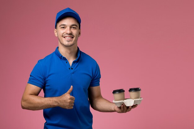 male courier in blue uniform holding delivery coffee cups and smiling on pink, uniform job worker service delivery