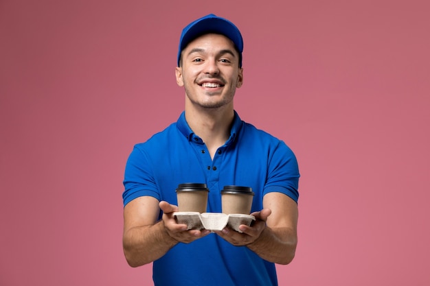 курьер-мужчина в синей униформе с улыбкой на розовом, держащий кофейные чашки с доставкой
