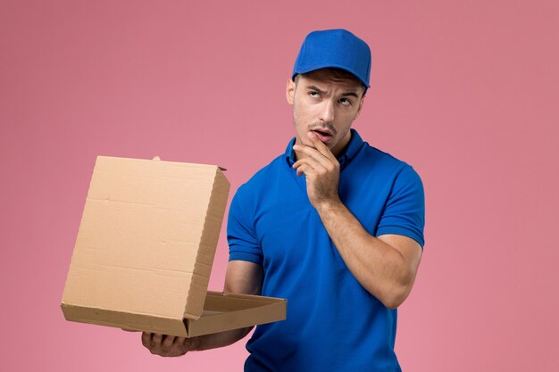male courier in blue uniform holding delivery food box opening it on pink, uniform service job delivery