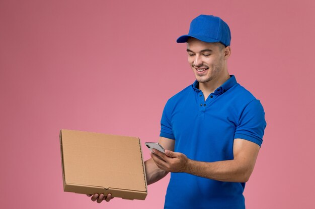 курьер-мужчина в синей форме держит коробку с доставкой еды и смартфон на розовом, униформе работник службы доставки
