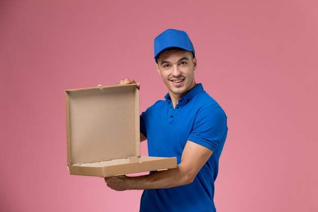 male courier in blue uniform holding and opening food box on pink, uniform service job delivery