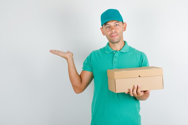 Male courier in green t-shirt with cap holding box and keeping opened hand