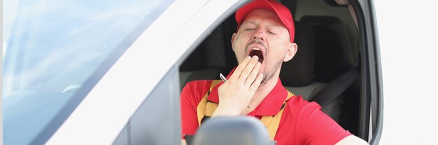 Male courier sitting behind wheel of car and yawning