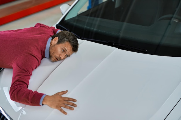 Male customer listening to car engine while leaning on a vehicle hood in a workshop