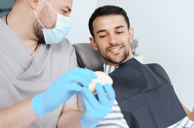 Male dentist in medical mask explaining artificial teeth to smiling patient in clinic