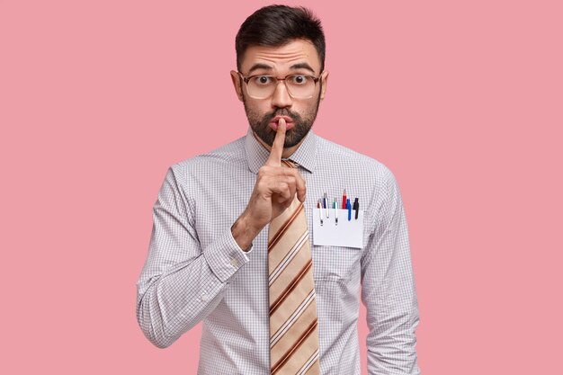 남성 디자이너는 공식적인 옷을 입고 입술에 손가락을 대고 셔츠 주머니에 연필과 펜이 든 빈 카드를 가지고 있습니다.