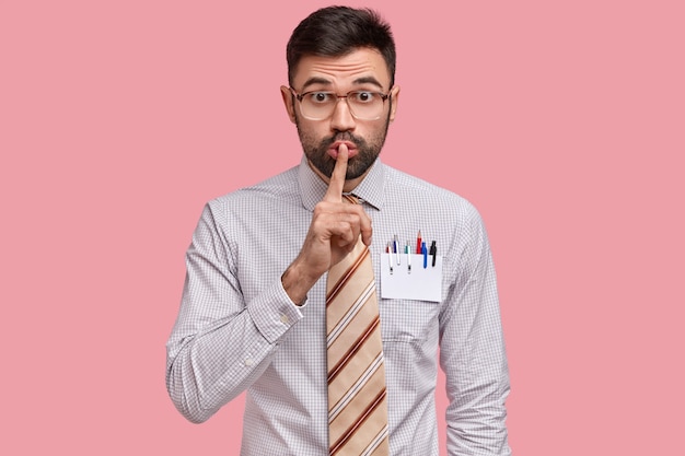 Мужчина-дизайнер держит палец на губах, одет в строгую одежду, имеет чистую карточку с карандашом и ручками в кармане рубашки.
