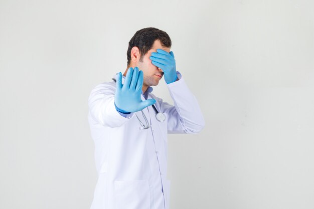 Male doctor doing stop gesture with palm on eyes in white coat, gloves and looking ashamed. front view.