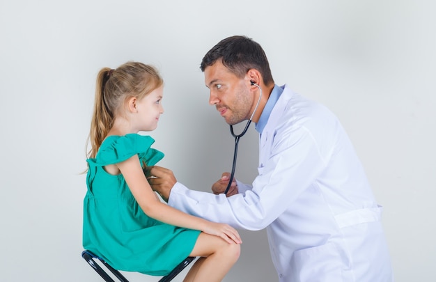 Male doctor examining little girl's heart with stethoscope in white uniform and looking glad