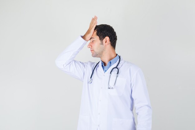 Male doctor holding hand on forehead in white coat and looking regretful