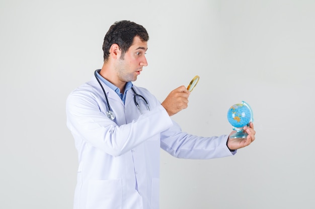 Male doctor holding magnifier over globe in white coat and looking surprised