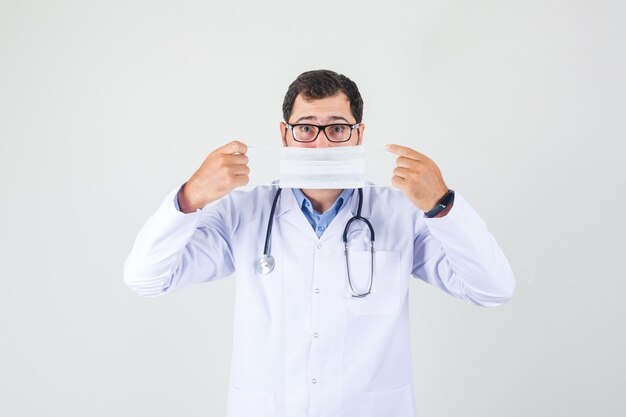 Male doctor holding medical mask over mouth in white coat, glasses and looking careful. front view.