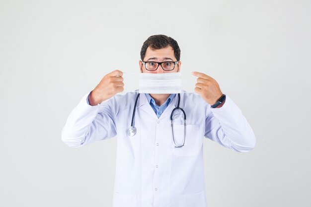 白衣、眼鏡をかけて口に医療用マスクをかざし、注意深く見ている男性医師。正面図。