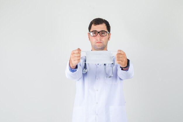 Male doctor holding medical mask in white coat, glasses and looking careful. front view.