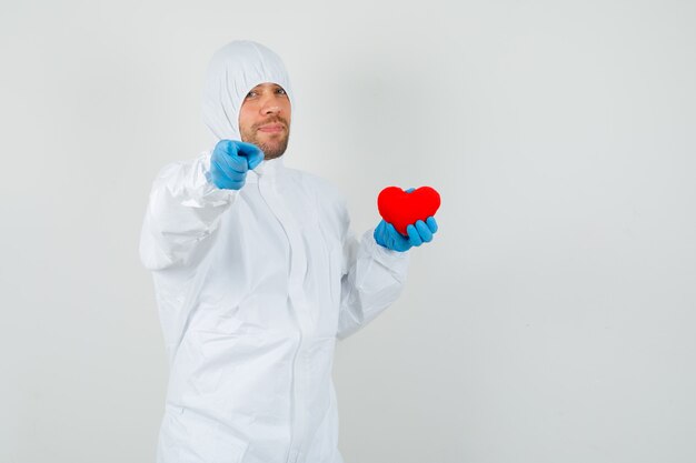 Male doctor holding red heart, pointing at camera in protective suit