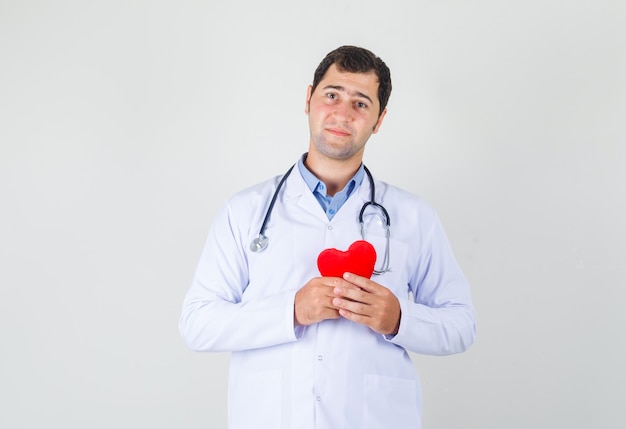 Мужчина-врач держит красное сердце в белом халате и смотрит с надеждой