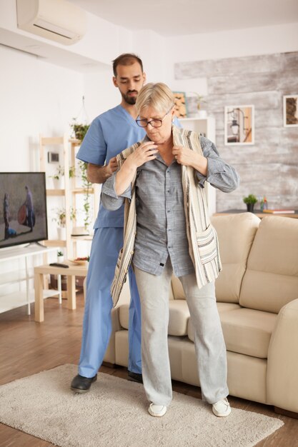 Male doctor in nursing home wearing blue uniform helping senior woman to dress.