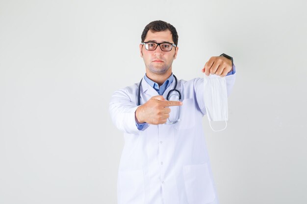 Male doctor pointing finger at medical mask in white coat, glasses front view.