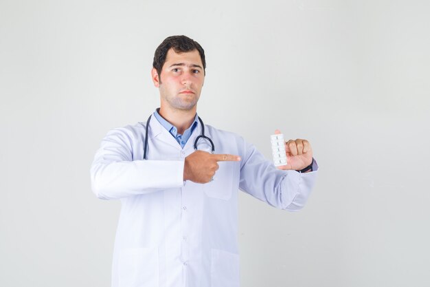 Male doctor pointing finger at pills in white coat and looking serious. front view.