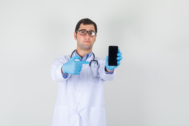 Male doctor pointing finger at smartphone in white coat, gloves, glasses and looking confused