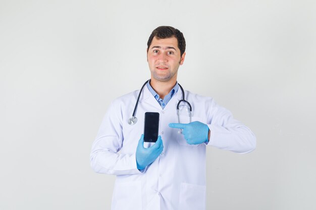 Мужской доктор указывая пальцем на смартфон в белом халате, перчатках