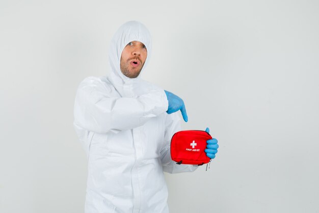 Male doctor pointing at first aid kit in protective suit, gloves and looking surprised.