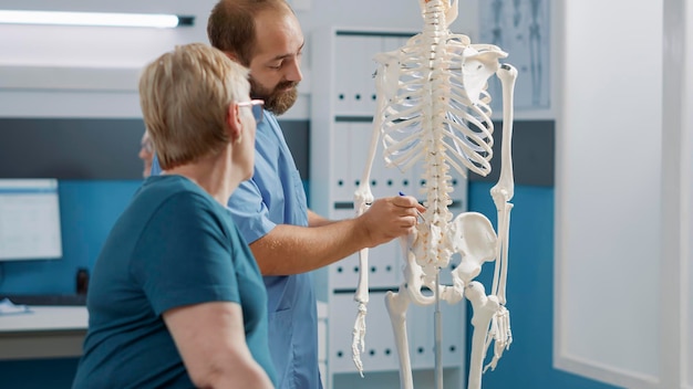 Male doctor pointing at human skeleton to show spinal cord and explain mechanical disorders in rehabilitation cabinet. Specialist explaining back bones system for physical therapy.