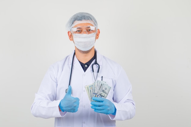 Male doctor in protective clothes holding dollar banknotes, making thumb up