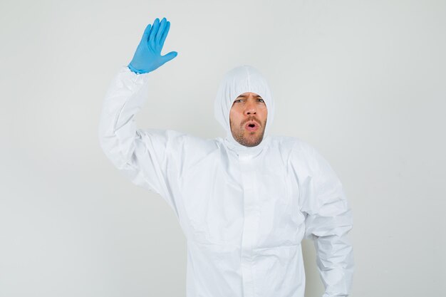 Мужчина-врач поднимает руку и ладонь в защитном костюме, перчатках и выглядит смущенным, вид спереди.