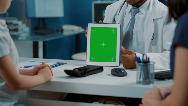 Врач-мужчина показывает шаблон зеленого экрана на планшете на приеме у врача. Практик держит пустой хроматический ключ с макетом фона и изолированным пространством для копирования на гаджете. Крупным планом