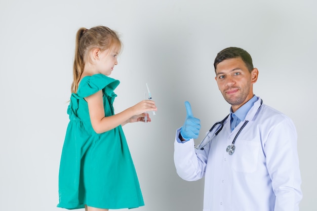 Male doctor showing thumb up while child holding syringes in white coat front view.