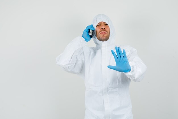 Male doctor talking on mobile phone with stop gesture in protective suit