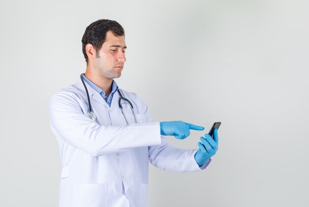 Мужчина-врач трогает смартфон пальцем в белом халате, перчатках и выглядит серьезно.