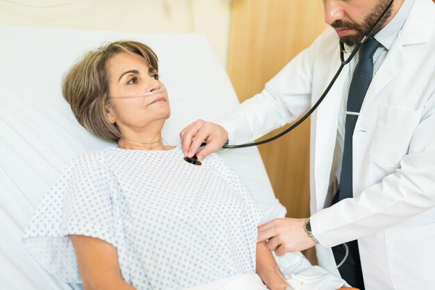 Male doctor treating senior patient with stethoscope on bed at hospital