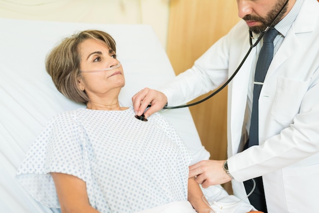 Male doctor treating senior patient with stethoscope on bed at hospital