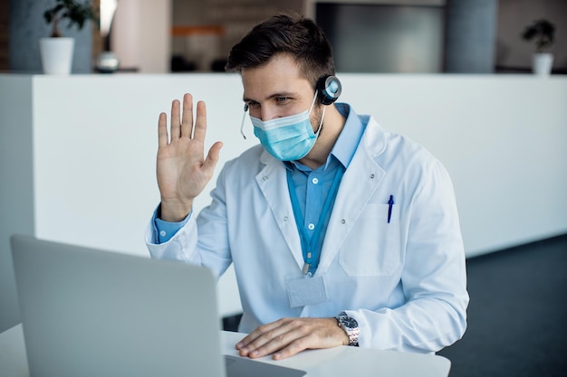 Врач-мужчина машет рукой во время видеозвонка через ноутбук в медицинской клинике
