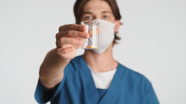 白い背景の上のカメラでガラスフラスコの丸薬を示す医療マスクを身に着けている男性医師病院で働くインターン