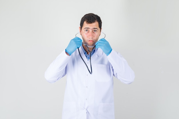 Male doctor wearing stethoscope in white coat, gloves and looking focused. front view.