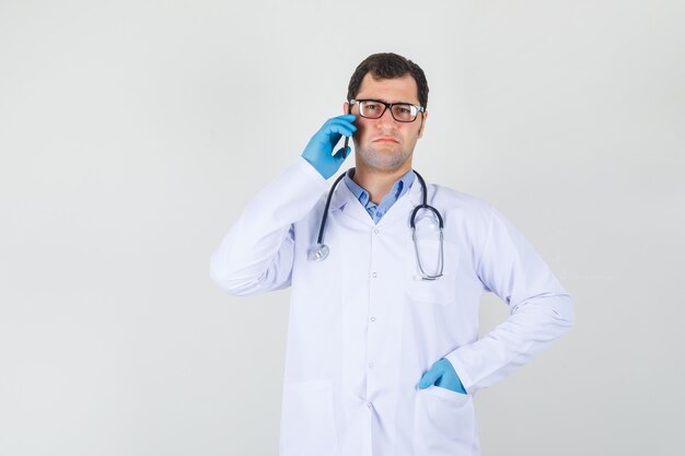 Мужчина-врач в белом халате, перчатках, очках разговаривает по телефону с рукой в кармане и выглядит недовольным