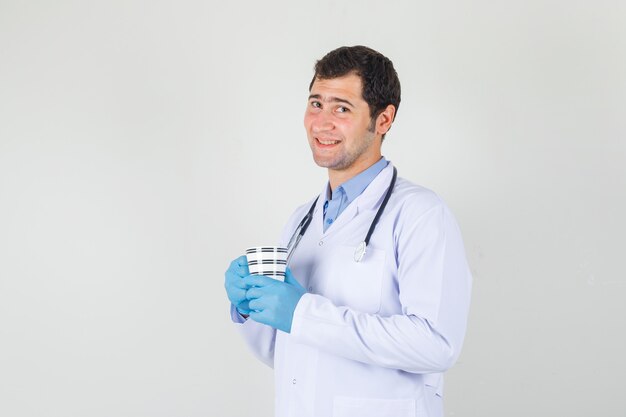 Male doctor in white coat, gloves holding cup of drink and smiling .