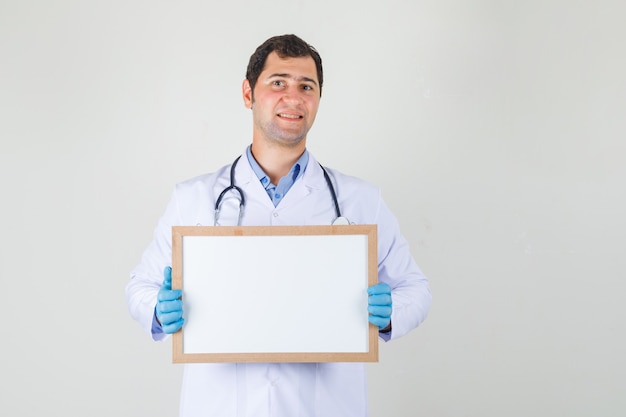 Male doctor in white coat, gloves holding white board and looking cheerful