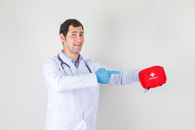 Medico maschio in camice bianco, guanti che indicano il dito al kit di pronto soccorso e che sembra allegro