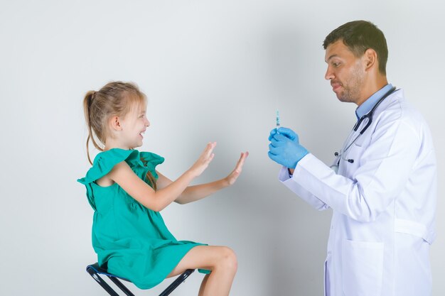 Male doctor in white uniform, gloves holding syringe while child gesturing no