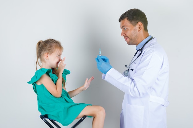 Male doctor in white uniform, gloves holding syringe while child looking with fear