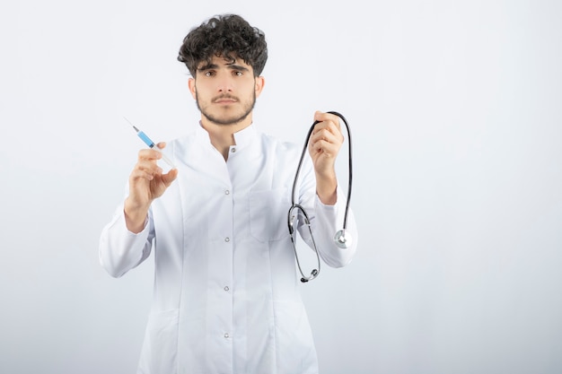 Male doctor in white uniform holding syringe and stethoscope in hands . 