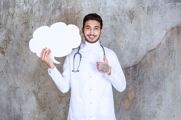 Male doctor with stethoscope holding a blank cloud shape info board and showing thumb up.