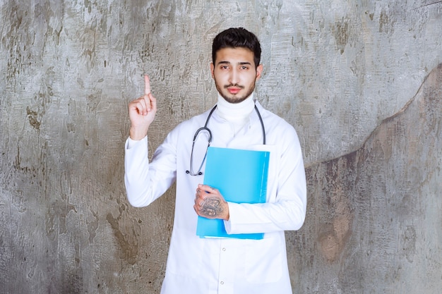 Врач-мужчина со стетоскопом держит синюю папку и поднимает руку, чтобы выразить беспокойство