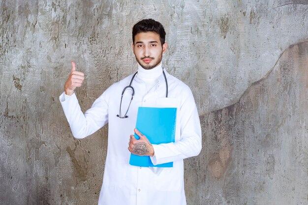 Medico maschio con lo stetoscopio che tiene una cartella blu e che mostra il segno positivo della mano.