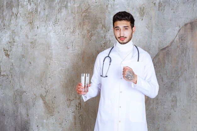 Мужчина-врач со стетоскопом держит стакан чистой воды и показывает положительный знак руки